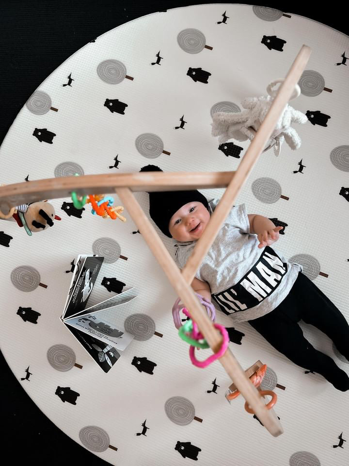 A baby lies on a round mat surrounded by colorful toys, smiling and reaching for them.