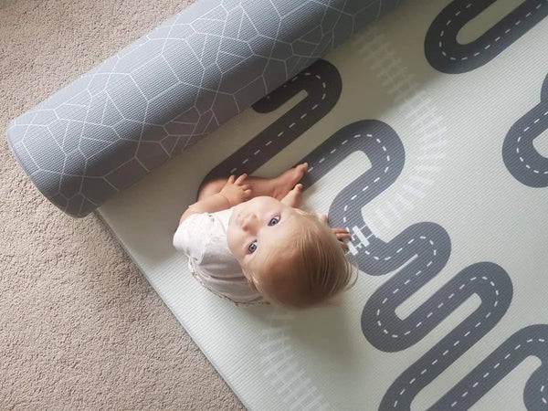 Kid sitting on Earl Grey Baby Driver playmat. Side A has a grey geometric pattern. Side B has a car track design.